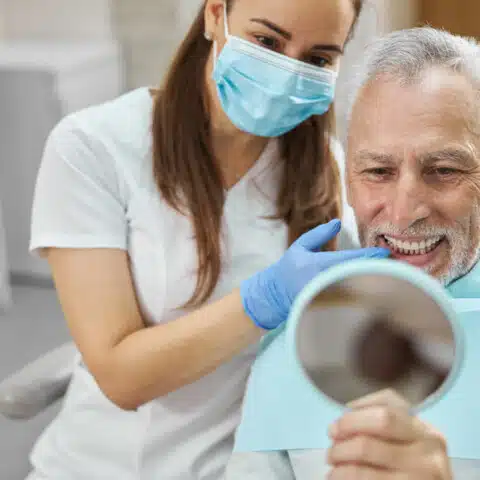 Older man looking at his teeth in a dental mirror with dentist